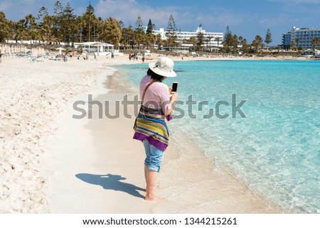 Barefoot woman in hat on the beach taking photos of the sea with smartphone. Summer holidays, vacation, travel and people concept