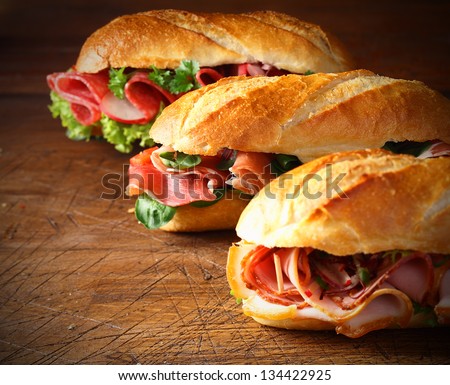 Assorted delicious baguette sandwiches filled with thinly sliced ham or salami and fresh green lettuce or basil arranged in an oblique row on an old wooden table with copyspace