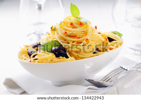 Close up shot of spaghetti pasta in a white bowl with olives on a white napkin with wine glasses in the background.