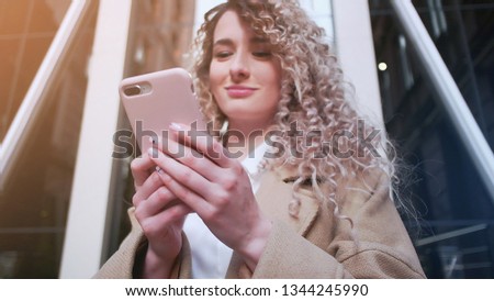 Beautiful young woman with blonde hair messaging on the smart-phone at the city street background.