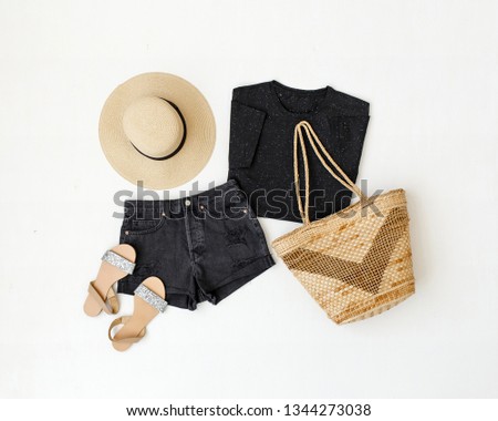 Black t-shirt, denim short, straw woven basket beach bag, boater hat, flat sandans on white background. Overhead view of woman's casual outfit. Trendy simple basic minimalistic look. Women clothes. Royalty-Free Stock Photo #1344273038