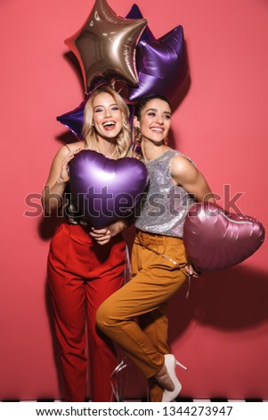 Image of two caucasian girls 20s in stylish outfit laughing and holding festive balloons isolated over red background
