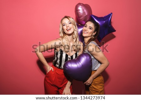 Image of two joyous girls 20s in stylish outfit laughing and holding festive balloons isolated over red background