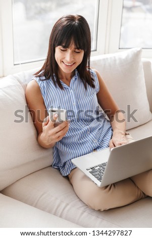 Portrait of middle-aged woman 40s in elegant clothes using cell phone and laptop while sitting on couch in bright apartment