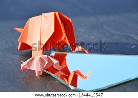 A small and a large origami elephant folded from red and pink paper stand on a monochrome surface in front of a mirror shard that looks like the two elephants are standing in front of a water hole