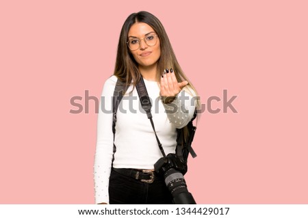 Young photographer woman inviting to come with hand. Happy that you came on isolated pink background