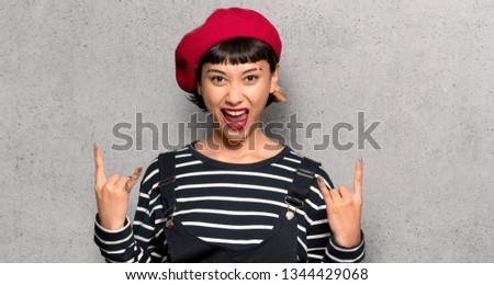 Young woman with beret making rock gesture over textured wall