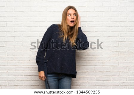 Blonde woman over brick wall listening to something by putting hand on the ear