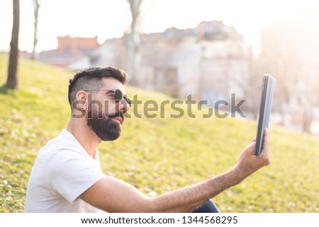 Hipster Young Man Taking a Selfie with his Mobile Phone in a Park - Using the Cellphone Outdoors in a Sunny Day of Summer. Lifestyle Concept.