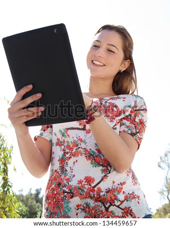 Portrait of a joyful young woman in a park using a digital tablet pad during a sunny day with a blue sky and smiling.