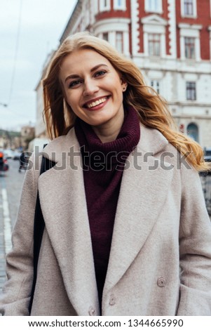 Beautiful young woman with blonde hair in stylish coat smiling at the camera while walking on the city street on spring day. Happy girl. City portrait