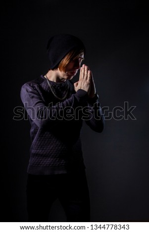 unusual freak young guy in black knit cap and red bangs stands in praying pose with hands folded front of face with eyes closed in studio with deep shadows and  black isolated background. copy space.