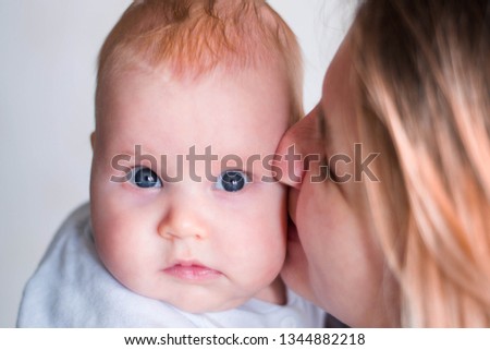 Mum kisses a baby baby in a cheek. The girl has a funny look. Caucasian child, blue eyes and red hair. Mom is not in focus.