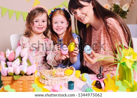 young woman mother with little girls daughters preparing for the celebration of Easter and coloring eggs sitting at the table on the background of the Easter decor