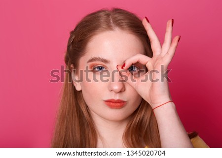 Studio shot of attractive serious young woman, makes ok sign, covers her eye, expresses confidence, model poses against pink background, being photographing in studio. People and gesture concept.