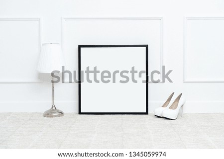 There are beautiful flowers and various objects near the rectangle blank picture frame in front of white wall on the carpet living room.
