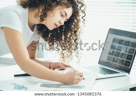 The curly woman at the table painting a picture near the laptop