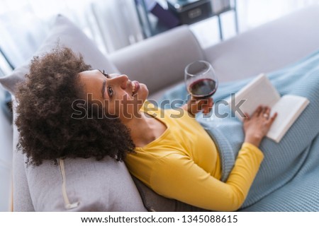 Young pretty woman sitting at opened window drinking wine and reading a book enjoys of rest. Unwinding the way she knows best. Enjoying a good literature Royalty-Free Stock Photo #1345088615
