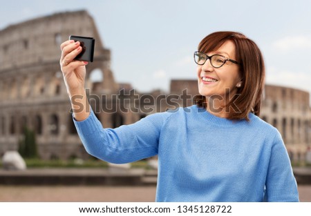 travel, tourism and technology concept - smiling senior woman in glasses taking selfie by smartphone over coliseum background