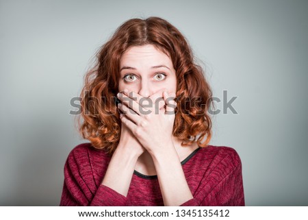 cute red-haired girl frightened and covers mouth with hands, isolated on gray background