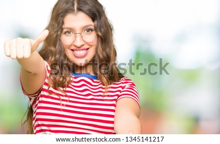Young beautiful woman wearing glasses approving doing positive gesture with hand, thumbs up smiling and happy for success. Looking at the camera, winner gesture.