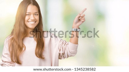 Young beautiful brunette woman wearing pink sweatshirt over isolated background with a big smile on face, pointing with hand and finger to the side looking at the camera.