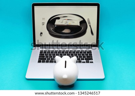 Small piggy bank standing on notebook and looking to laptop monitor,on the computer screen picture on black plate dollars money with fork and knife isolated on blue background