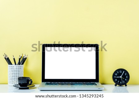 Front view of creative designer desktop with blank screen laptop, supplies and other gadgets on yellow wal.Mock up