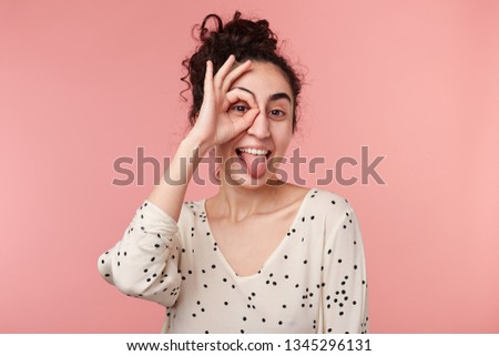 Portrait of funny attractive positive charming brunette girl with collected wave hair making okey symbol close to eye with one hand, shows tongue, in blouse with polka dots,isolated on pink background