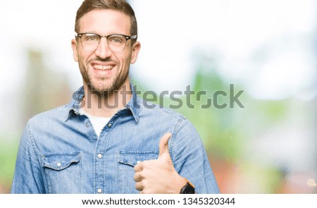 Handsome man wearing glasses doing happy thumbs up gesture with hand. Approving expression looking at the camera showing success.