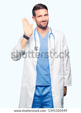 Handsome doctor man wearing medical uniform over isolated background Waiving saying hello happy and smiling, friendly welcome gesture