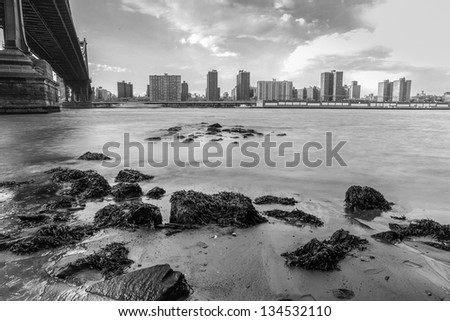 black and white view on manhattan skyline from under the brooklyn bridge with waves and rocks dumbo