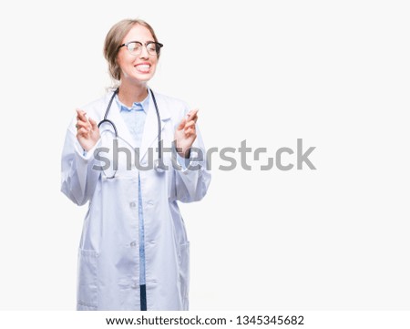 Beautiful young blonde doctor woman wearing medical uniform over isolated background smiling crossing fingers with hope and eyes closed. Luck and superstitious concept.