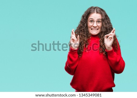 Beautiful brunette curly hair young girl wearing glasses and winter sweater over isolated background smiling crossing fingers with hope and eyes closed. Luck and superstitious concept.