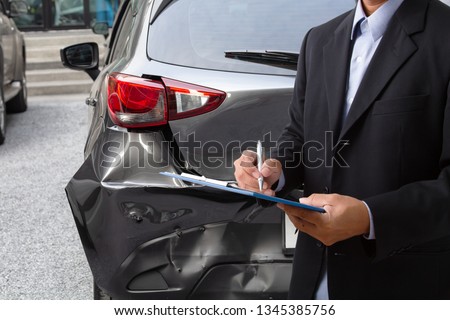 Side view of insurance officer writing on clipboard while insurance agent examining black car after accident. Royalty-Free Stock Photo #1345385756