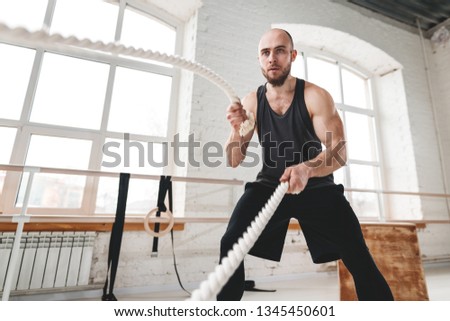 Strong man workout with battle ropes at light gym. Muscular sportsman doing cross excursion with ropes in workout gym