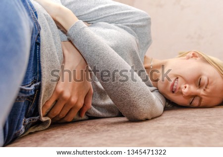 Blonde girl lying on the couch in blue jeans and a gray sweater, feels a pain in her stomach Royalty-Free Stock Photo #1345471322
