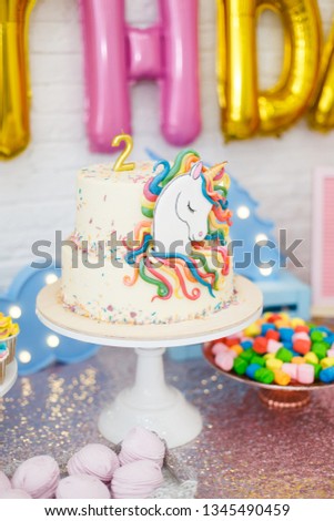 Room decoration for children's birthday with a unicorn. A table with sweets stands on the background of white brick walls: marshmallows, cupcakes, blueberry cake with chocolate cream, garland, cookie