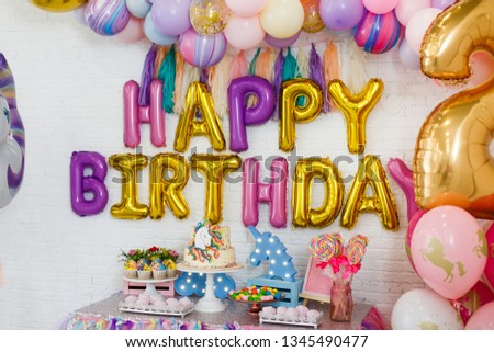 Room decoration for children's birthday with a unicorn. A table with sweets stands on the background of white brick walls: marshmallows, cupcakes, blueberry cake with chocolate cream, garland, cookie