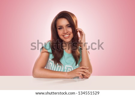 Smiling attractive young Latino woman sitting at a table facing the camera against a pink studio background with highlight