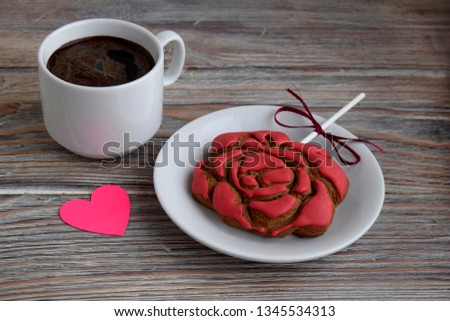 Gingerbread in the form of a flower of a red rose is on a white plate. Paper in the form of pink heart; and a cup with coffee are on a wooden table