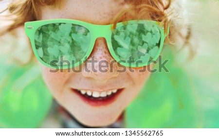 Close up portrait of smiling caucasian boy with freckles in green sunglasses, mirror reflection in glasses is  bokeh with hearts, green clothes, red hair.