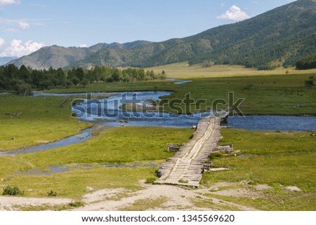 wooden bridge over a flowing blue river in a green valley of the Altai mountains with trees and mountains