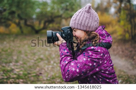 A young girl in a purple jacket and hat walks with a camera and takes pictures of nature in the fall. Baby photographer. Happy childhood. Portrait and photography.