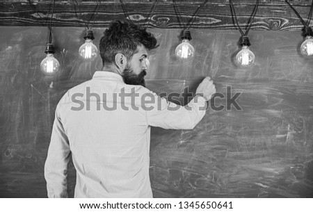 Man with beard stand in front of chalkboard, rear view. Guy busy with writing on chalkboard surface. Bearded hipster in shirt writing on chalkboard, copy space. Announcement concept.