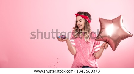Funny girl in a pink t-shirt with balloons and confetti gives a smile and emotions on a pink background. The concept of the party and holiday