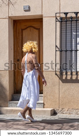 Butch man with hairy back, arms and legs totters along a street dressed as a look alike Hollywood actress in blond curly wig, flowing white dress and court shoes: fancy dress for a gay pride parade.