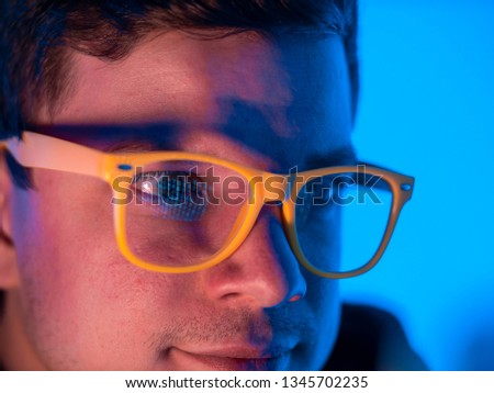 Close-up: A young hacker guy in yellow glasses is working on a cyber-attack computer, while the green symbols of a binary code are reflected in the glasses of his glasses. Programmer surprised