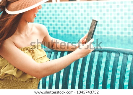 A beautiful girl wearing a hat, sitting on a chair, holding a phone, taking pictures at the pool side on holiday