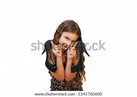 Emotional portrait of a cute little girl with long hair in a dress and with makeup that holds and tries on high-heeled shoes, isolated on a white background